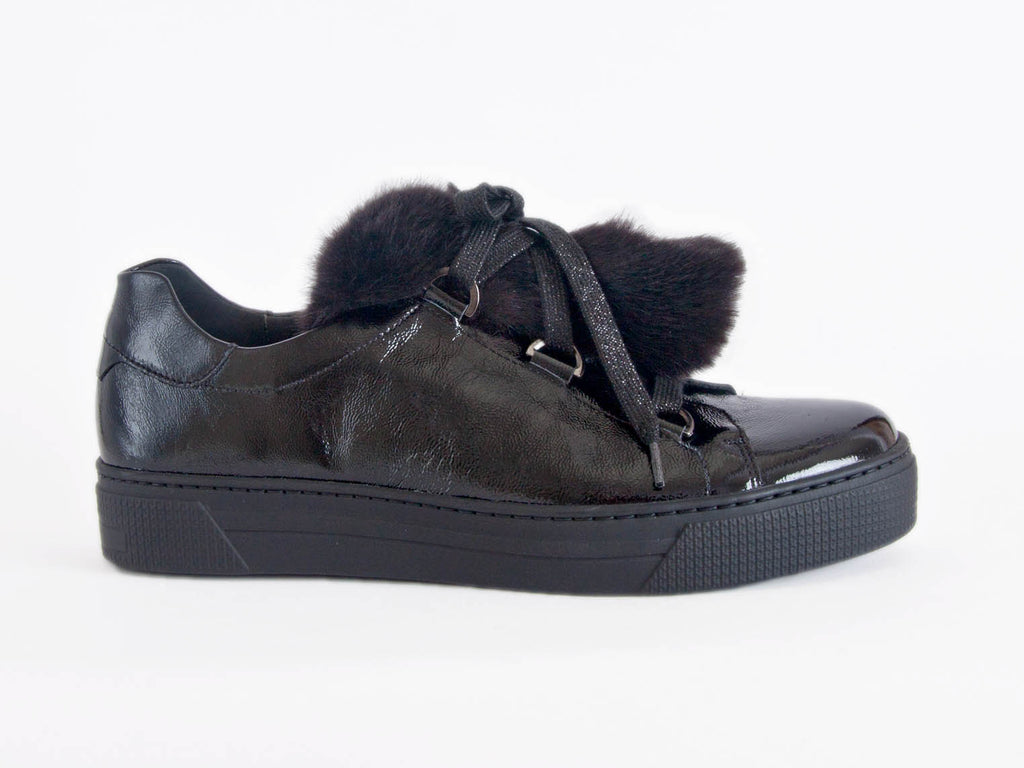 Side view of black patent leather trainer shoe with distinctive big fur tongue on top, slightly sparkly laces and thick flat black sole